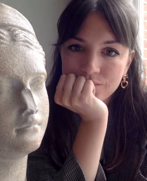A dark-haired woman in a plaid blazer rests her chin on her hand, looking at the camera, next to a classical stone bust.
