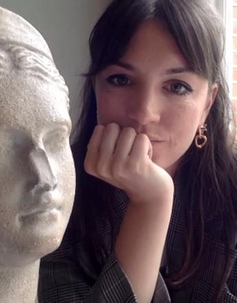 A dark-haired woman in a plaid blazer rests her chin on her hand, looking at the camera, next to a classical stone bust.