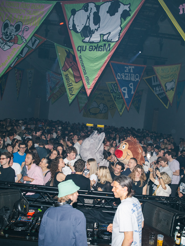 A crowded indoor party scene with DJs at a booth, a dense crowd of people, and colorful triangular banners featuring cartoon animals hanging from the ceiling.