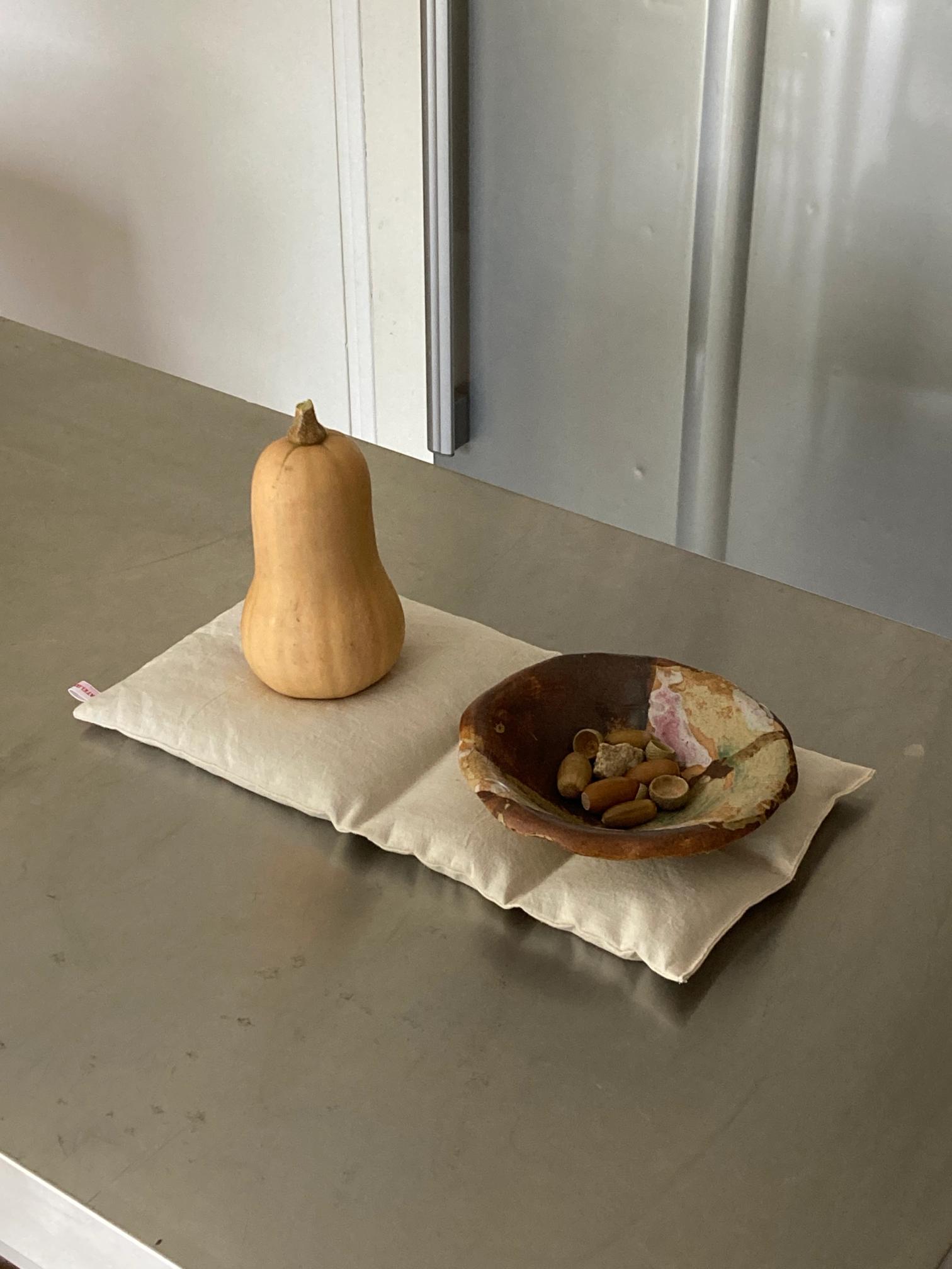 A butternut squash and a bowl of acorns rest on a light linen mat on a stainless steel counter.
