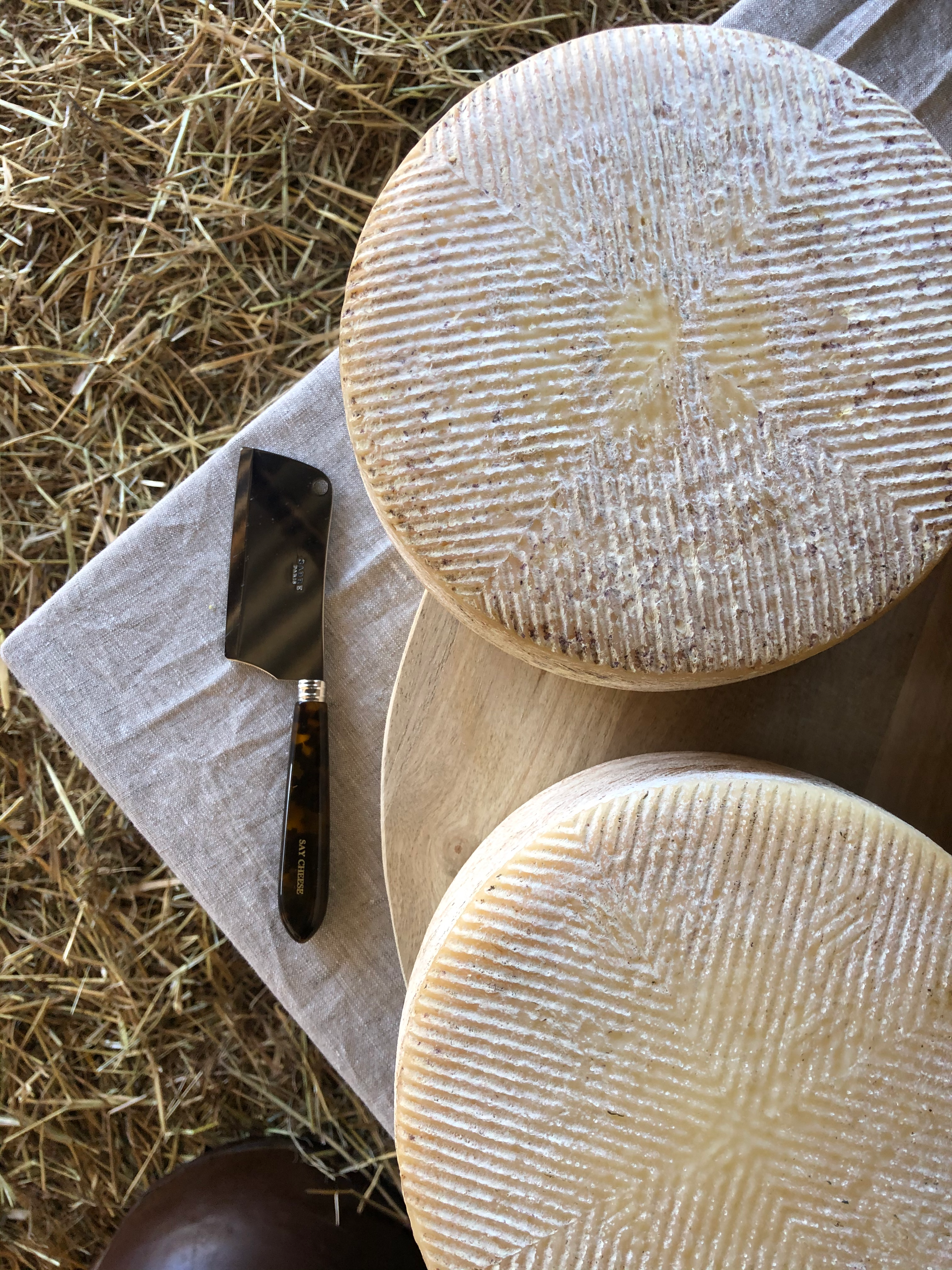Two round cheeses with ridged rinds, a small cleaver on a linen cloth, and a wooden board on hay.