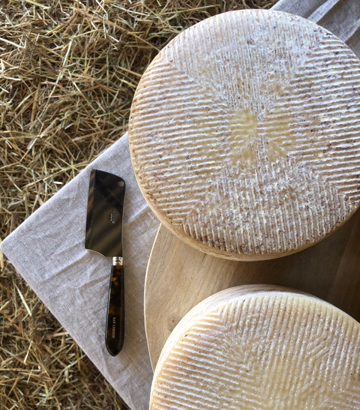 Two round cheeses with ridged rinds, a small cleaver on a linen cloth, and a wooden board on hay.