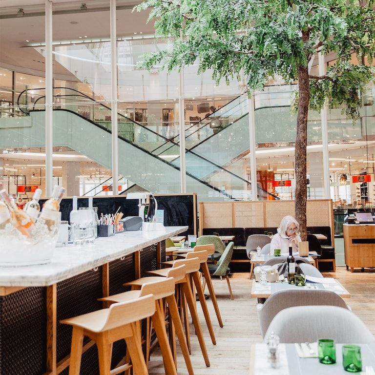 A bright, modern restaurant and bar with a large indoor tree, dining tables, and a woman seated, with escalators in the background.