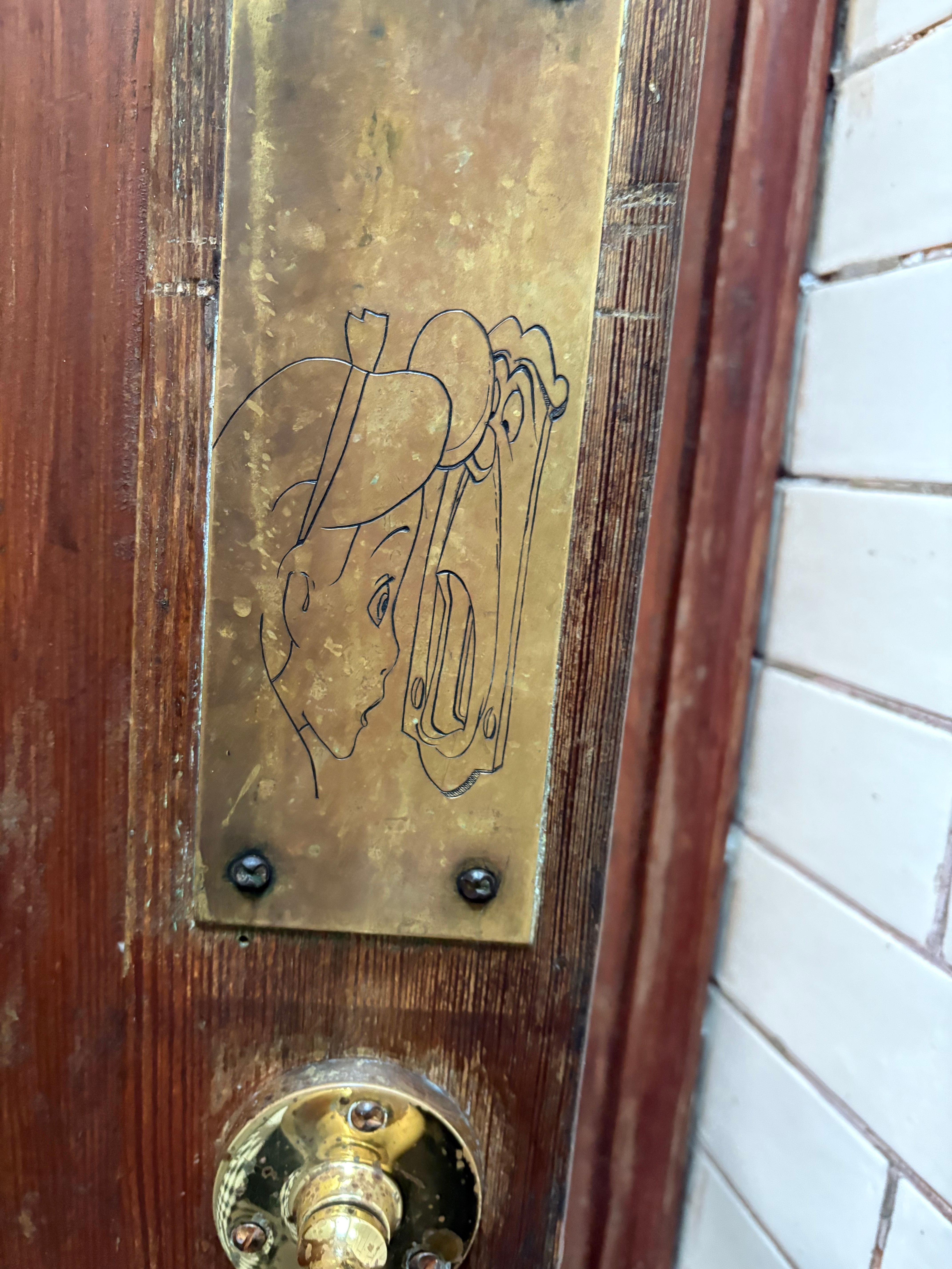 A brass plaque on a wooden door depicts Peter Pan peeking through a keyhole, with a brass doorknob below.