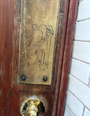 A brass plaque on a wooden door depicts Peter Pan peeking through a keyhole, with a brass doorknob below.