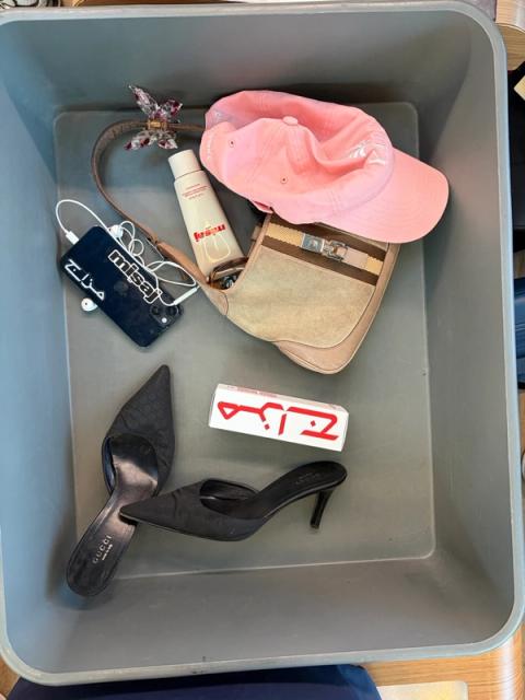 A grey security tray containing black high heels, a beige shoulder bag, a pink baseball cap, a phone, a lotion bottle, and a white box.