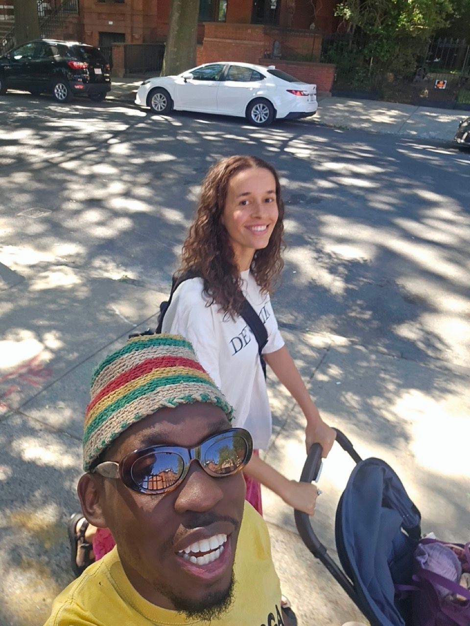 A smiling man in a colorful striped hat and sunglasses takes a selfie with a smiling woman pushing a stroller on a sunny street.