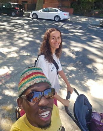 A smiling man in a colorful striped hat and sunglasses takes a selfie with a smiling woman pushing a stroller on a sunny street.