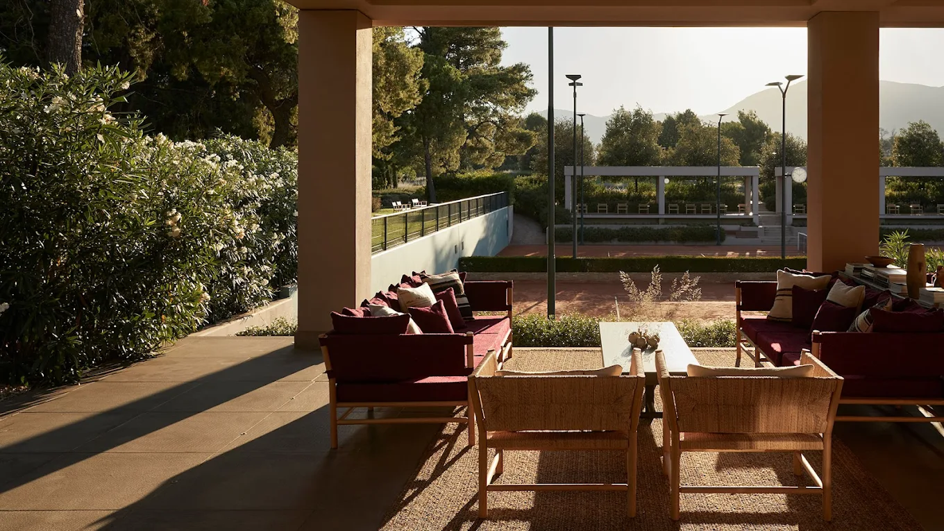 A sunny outdoor patio with comfortable seating overlooking a red clay tennis court, trees, and distant mountains.