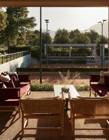 A sunny outdoor patio with comfortable seating overlooking a red clay tennis court, trees, and distant mountains.