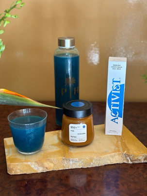 A glass of shimmering blue drink, a bottle of blue liquid, a jar of raw Manuka honey, and a white box for Activist Manuka Honey Mask on a light stone slab.