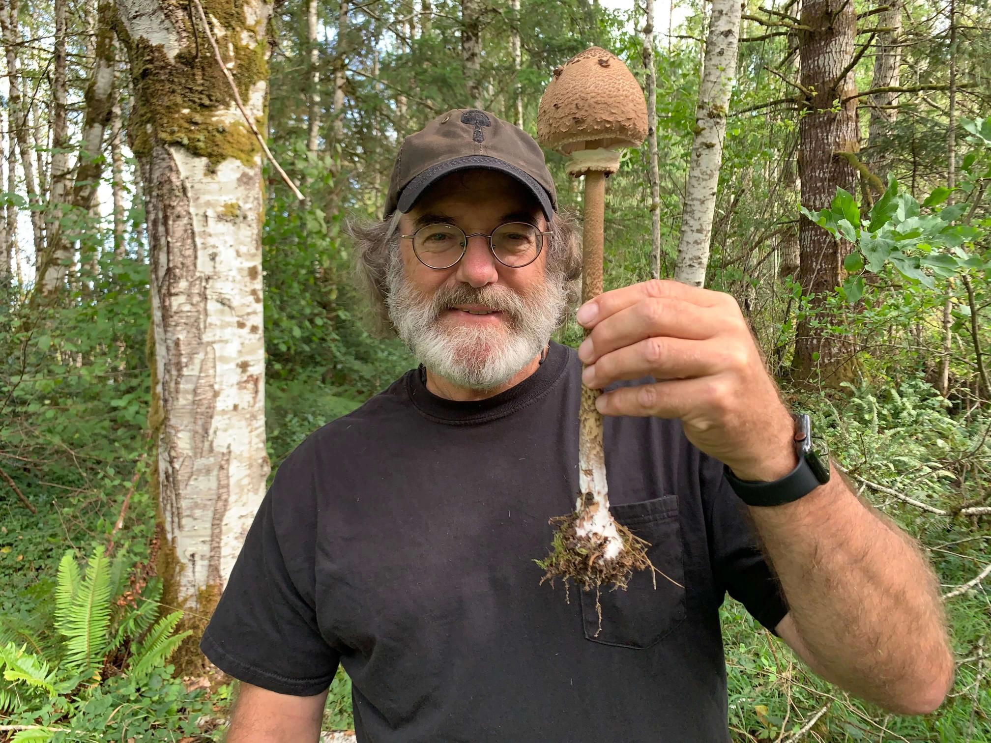 A smiling man with a beard and glasses holds a tall, dirt-covered mushroom in a forest.