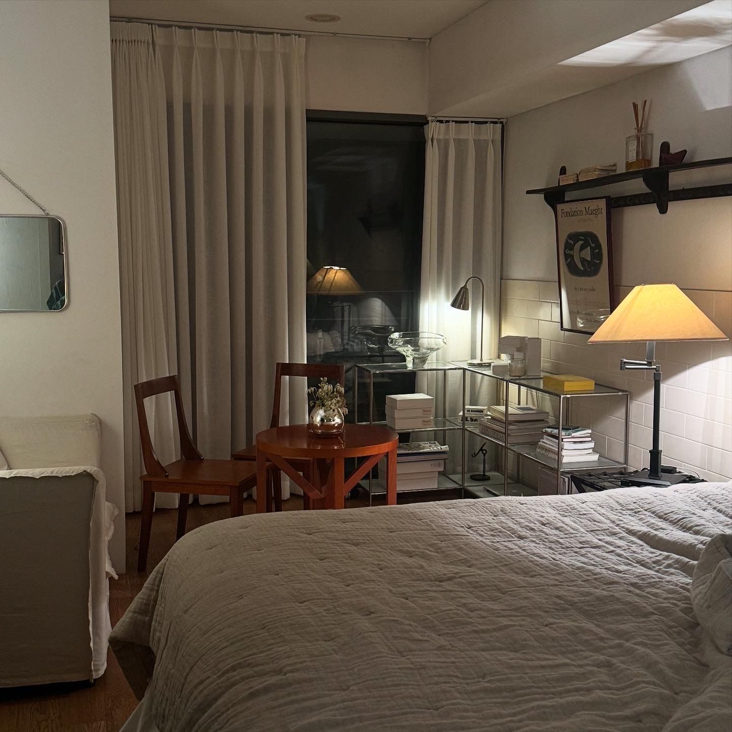 A cozy, dimly lit bedroom with a bed, a small table and chairs, a shelving unit, and a large window with sheer curtains, all illuminated by warm lamps.
