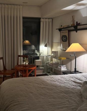 A cozy, dimly lit bedroom with a bed, a small table and chairs, a shelving unit, and a large window with sheer curtains, all illuminated by warm lamps.
