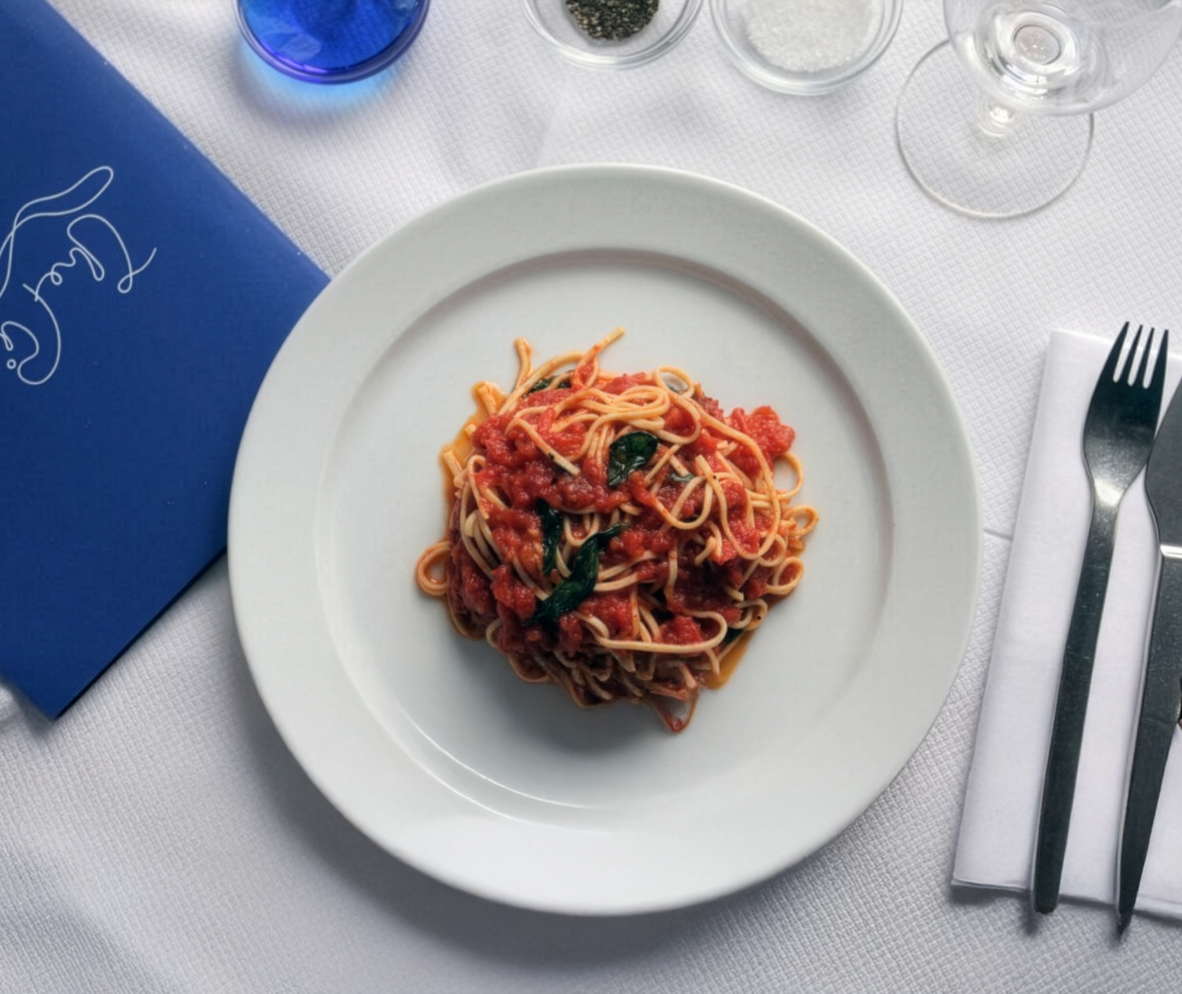 A plate of spaghetti with tomato sauce and basil on a white table setting.