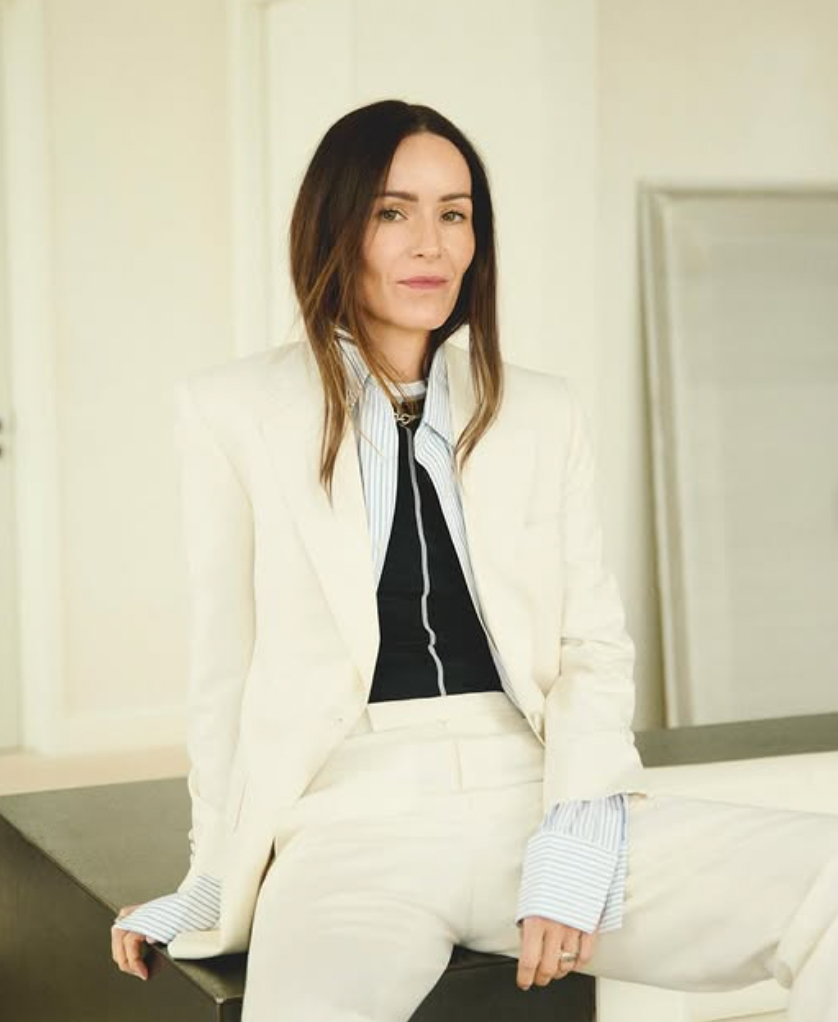Woman with long dark hair wearing a cream pantsuit and striped shirt, looking forward.