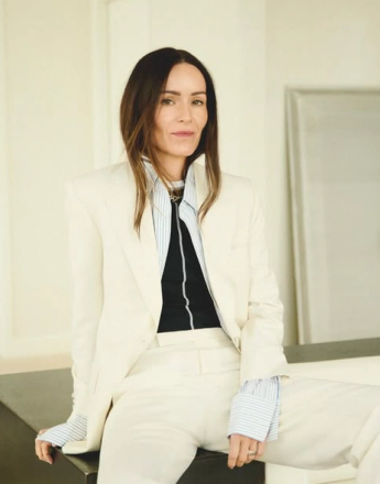 Woman with long dark hair wearing a cream pantsuit and striped shirt, looking forward.