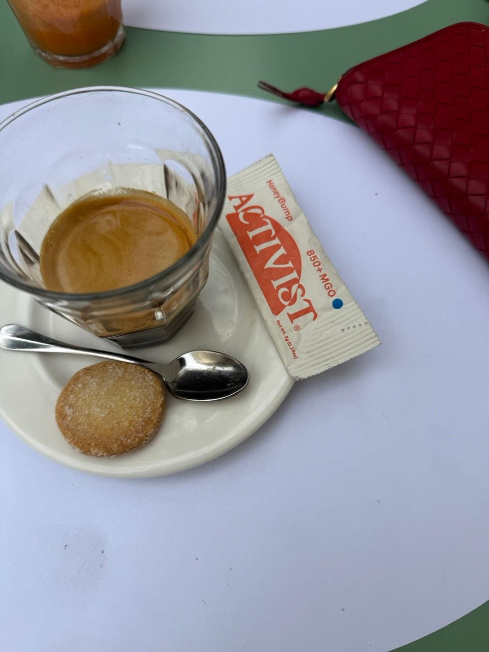 A glass of espresso with a cookie, spoon, and honey packet on a saucer next to a red wallet.