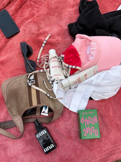 Beach essentials including a brown bag, phone, sunglasses, bikini, pink hat, sunscreens, and a green box, laid on a pink towel.