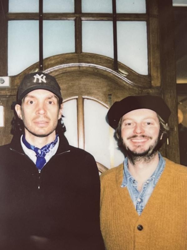 Two smiling men, one in a Yankees cap and blue bandana, the other in a beret and yellow cardigan.