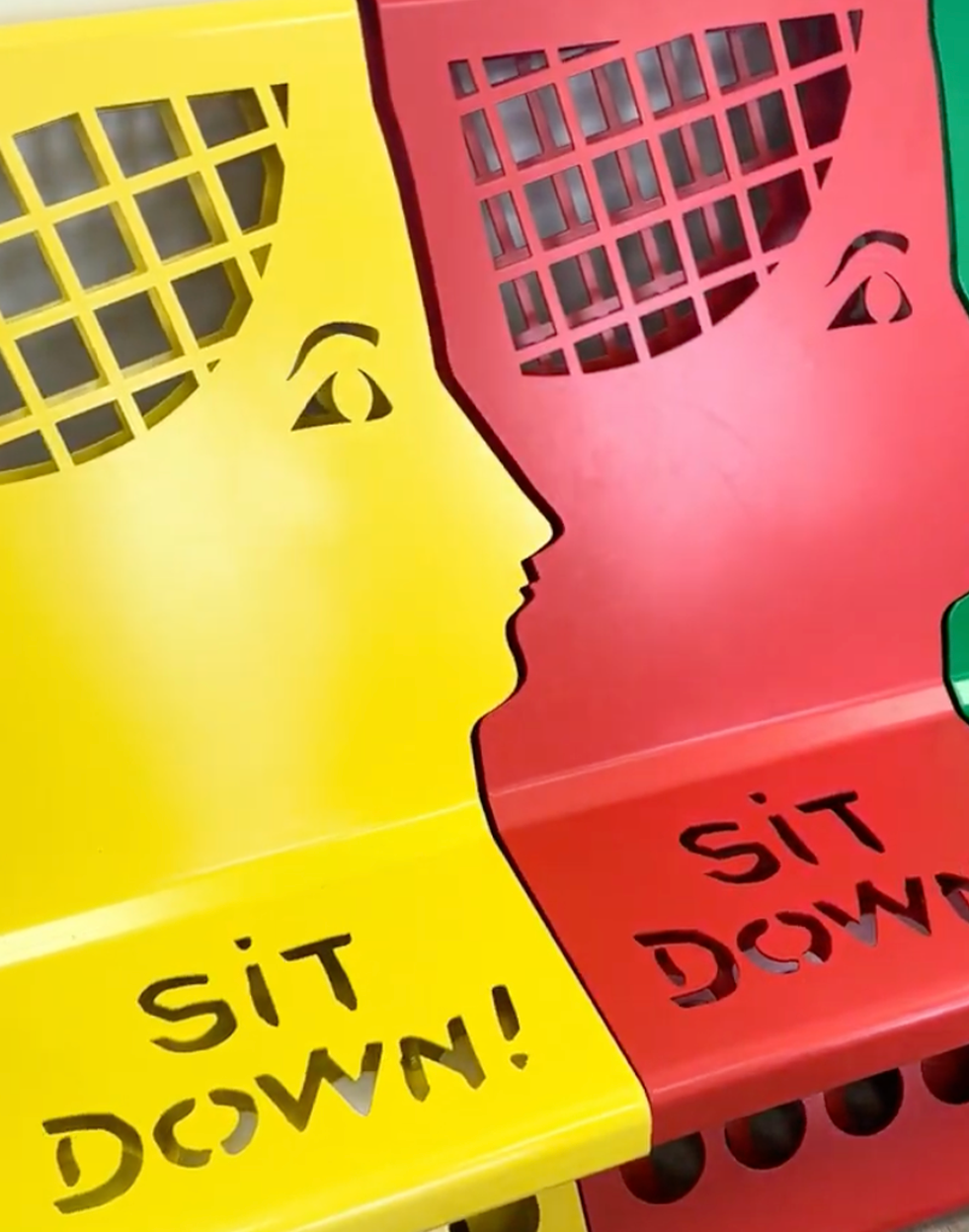 Yellow and red chairs, shaped like human profiles with grid backs, face each other, with "SIT DOWN!" cut into their seats.