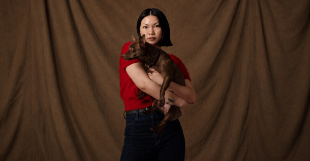 An Asian woman in a red top holds a small brown dog against a brown draped background.