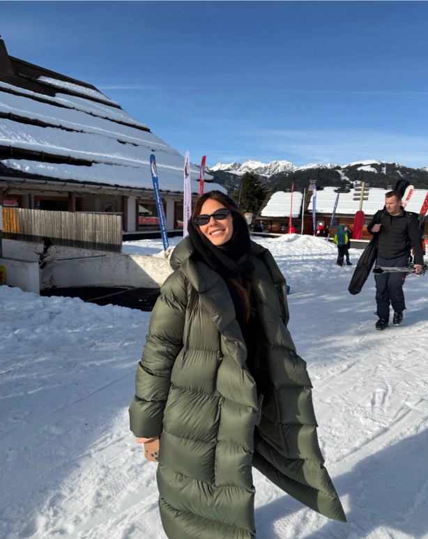 A smiling woman in a long green puffer coat and sunglasses stands in a snowy ski resort.