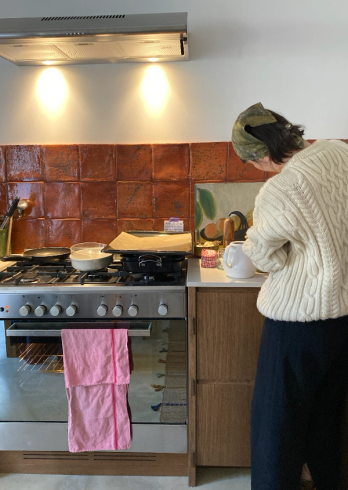 A person wearing a cream cable-knit sweater and a green headscarf prepares food at a stove in a kitchen with orange tiled walls.
