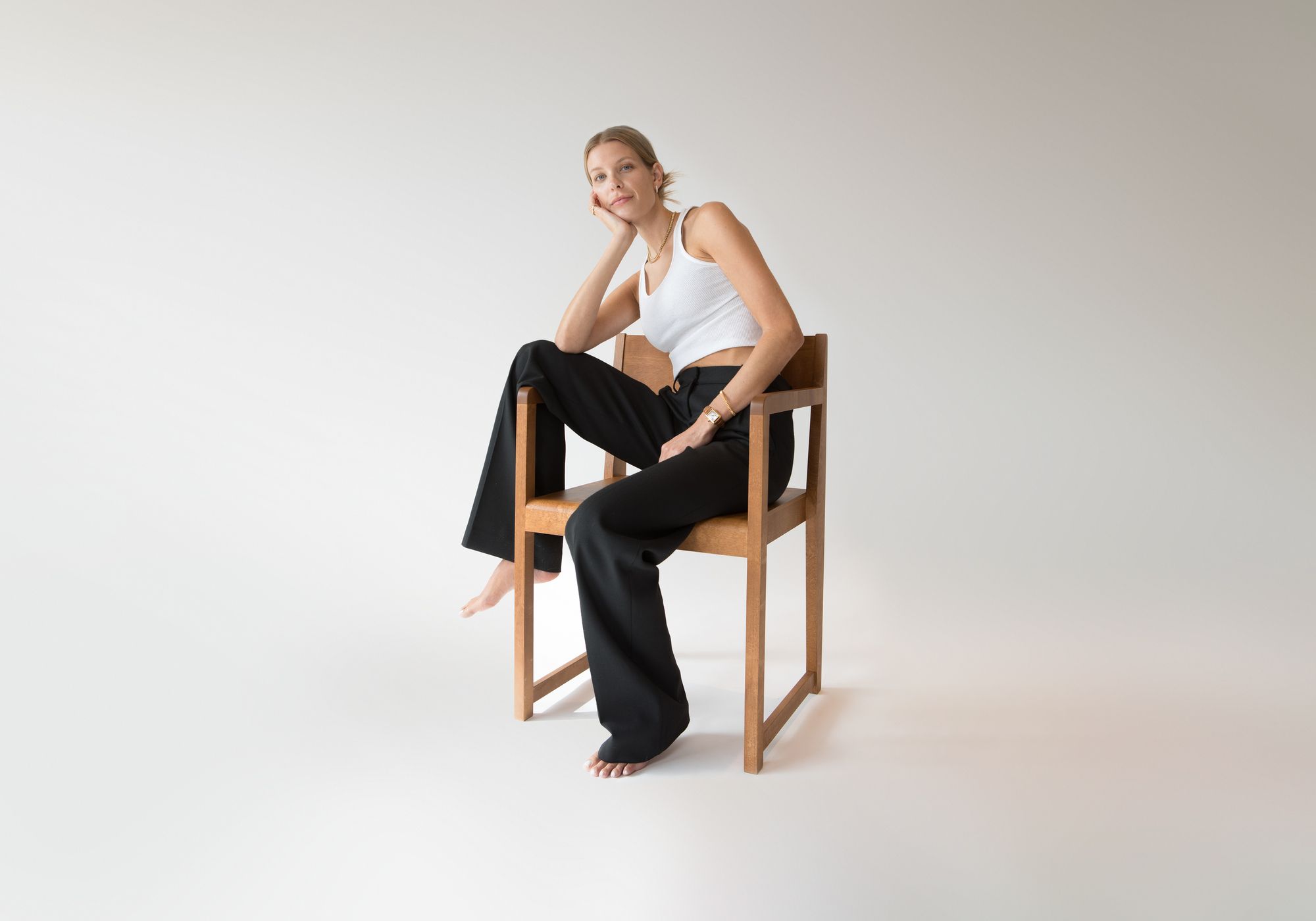 A blonde woman in a white tank top and black pants sits barefoot on a wooden chair, head in hand, looking at the camera.