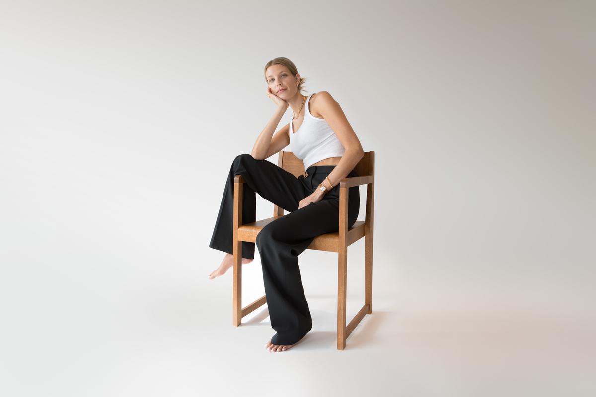 A blonde woman in a white tank top and black pants sits barefoot on a wooden chair, head in hand, looking at the camera.