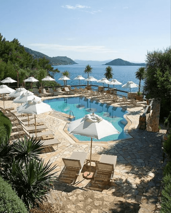 Outdoor swimming pool with white umbrellas and sun loungers, overlooking a clear blue sea and distant islands.