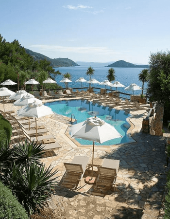 Outdoor swimming pool with white umbrellas and sun loungers, overlooking a clear blue sea and distant islands.