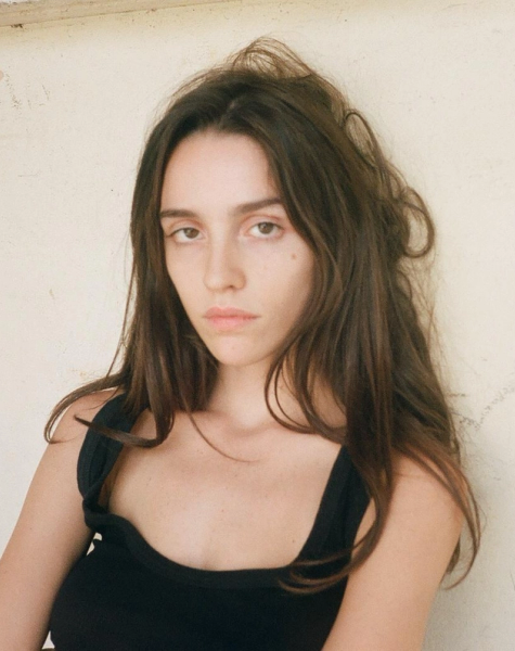 A young woman with long brown hair and a black top looks directly at the camera against a light wall.