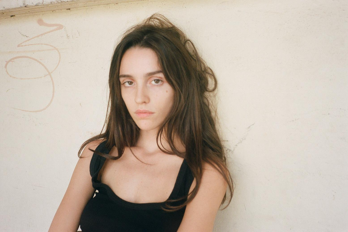 A young woman with long brown hair and a black top looks directly at the camera against a light wall.