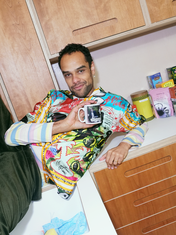 A smiling man in a colorful, graphic shirt holds a mug while leaning on a wooden cabinet, with tea items on a shelf behind him.