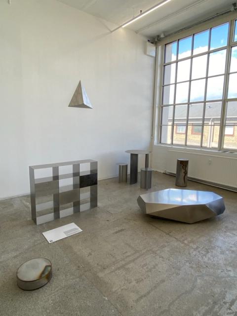 A modern gallery displays metallic, geometric sculptures, including a hanging pyramid, a checkered block, and a faceted table, on a concrete floor.