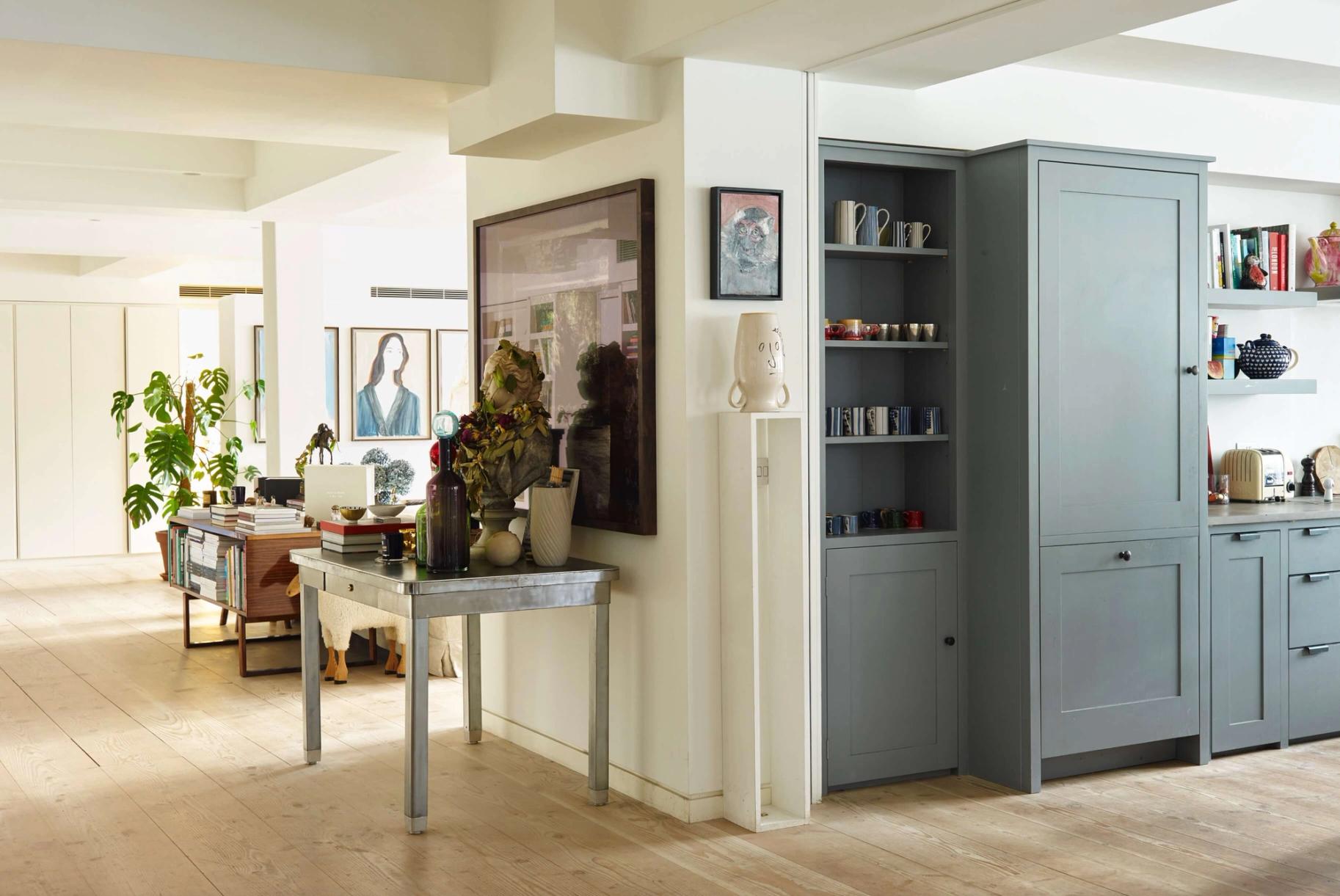 An open-plan room with a light wood floor, a stylish living area on the left with a console table and artwork, and a modern kitchen on the right with light blue-grey cabinets.