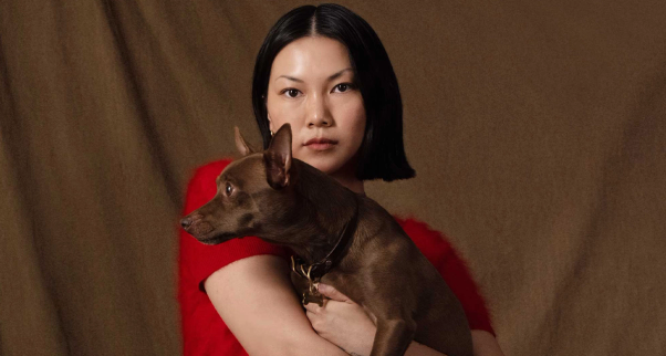 A woman with short dark hair in a red top holds a small brown dog.