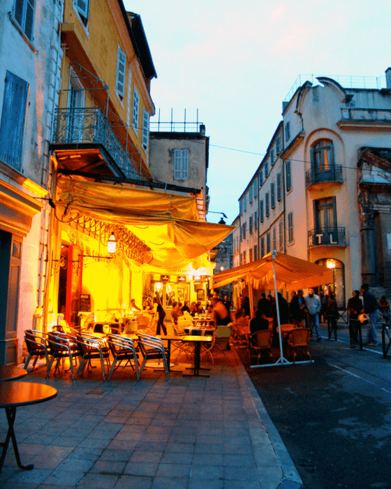 A warmly lit outdoor cafe with yellow awnings and diners on a narrow street at twilight.