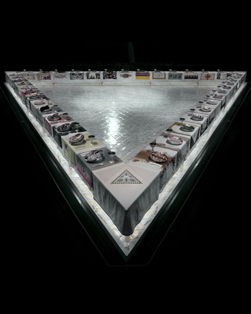 A triangular art installation, dramatically lit from below, featuring a large dinner table with individual place settings and textile art, including a pyramid and eye motif at the apex, against a dark background.