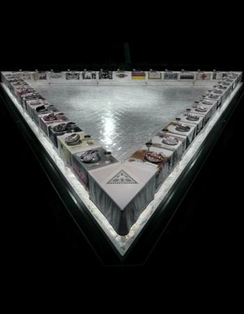 A triangular art installation, dramatically lit from below, featuring a large dinner table with individual place settings and textile art, including a pyramid and eye motif at the apex, against a dark background.
