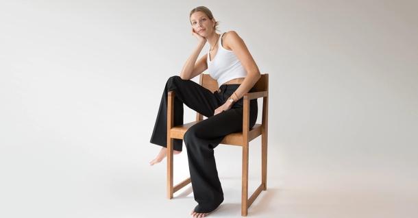 A barefoot woman in a white crop top and black wide-leg pants sits casually on a wooden chair against a white background.