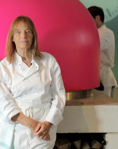 A woman in a chef's uniform stands in front of a bright pink pizza oven with a fire burning inside.