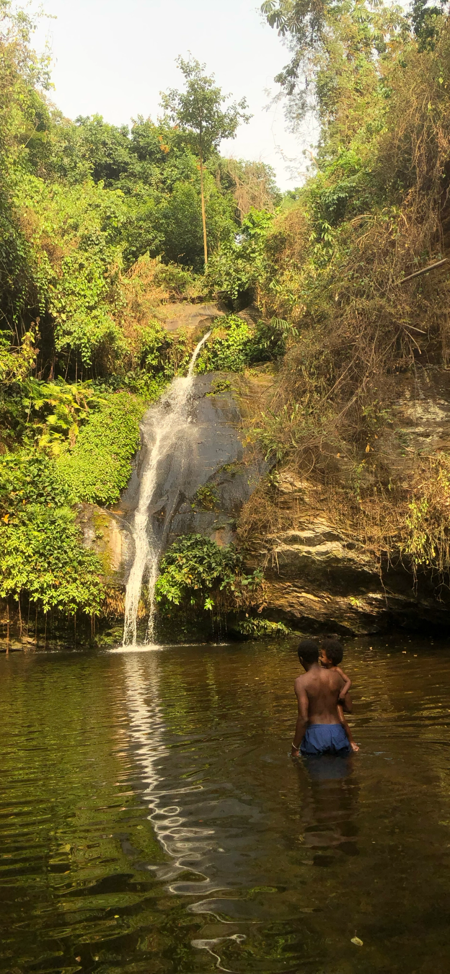 A person carrying a child on their back wades in a pool at the base of a waterfall, surrounded by dense jungle.
