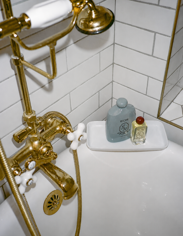 White bathtub with polished gold faucet and showerhead against white subway tiles, holding two bottles on a tray.