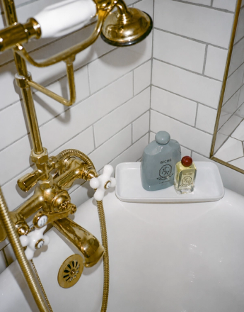 White bathtub with polished gold faucet and showerhead against white subway tiles, holding two bottles on a tray.