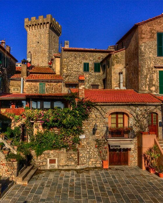 A cluster of stone buildings with red tile roofs and a tall medieval tower under a clear blue sky, overlooking a paved courtyard.