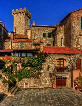 A cluster of stone buildings with red tile roofs and a tall medieval tower under a clear blue sky, overlooking a paved courtyard.