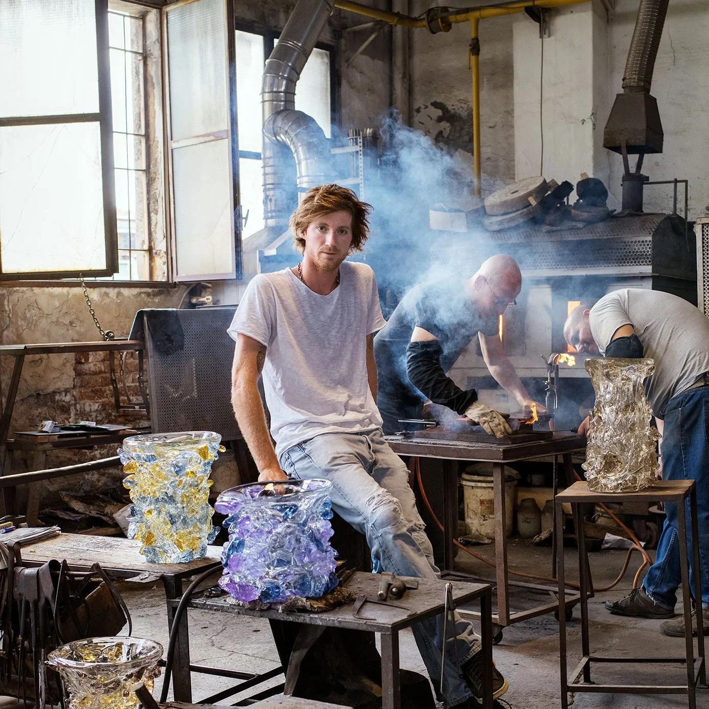 Man in a glassblowing studio with two textured glass vases; another artisan works with molten glass.