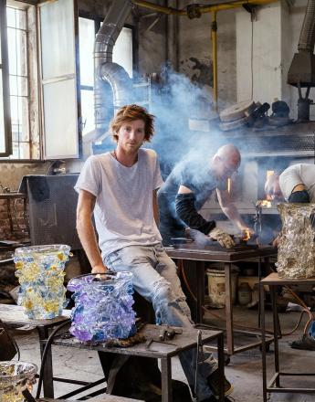 Man in a glassblowing studio with two textured glass vases; another artisan works with molten glass.